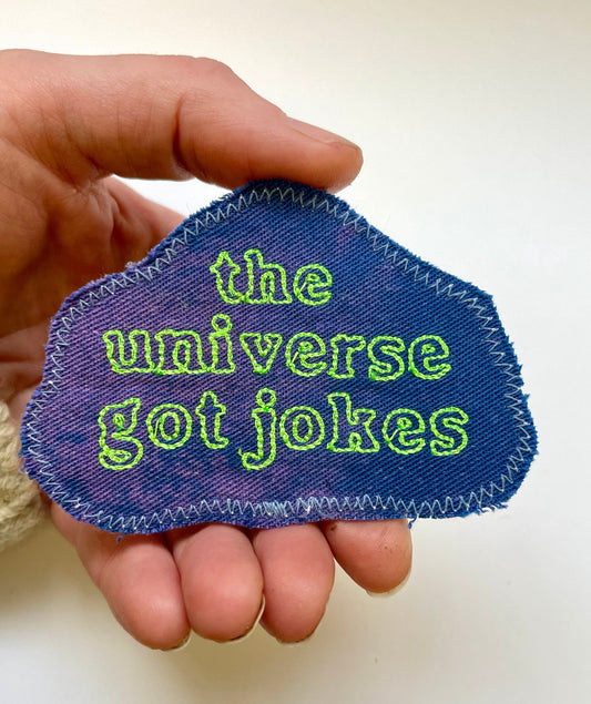 a hand holding a patch that says the universe got jokes